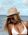 Person sitting at the beach wearing the Anya & Niki Essential Hat - a natural colored crochet straw fedora hat with ivory rope band.