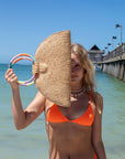 Person holding natural raffia straw half-moon clutch with rainbow colored wrapped circle handle and leather sides.