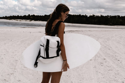 Person at beach with surf board, holding the Brooker white mesh backpack with black trim details.