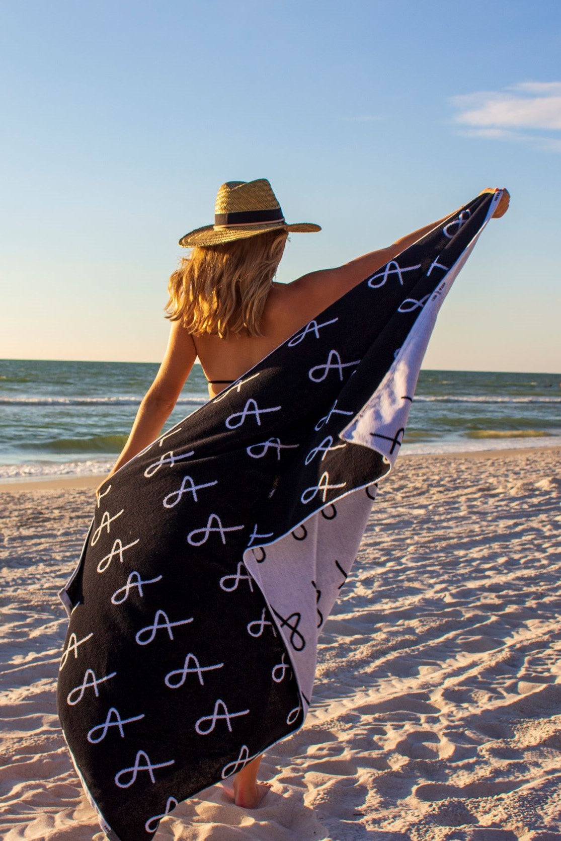 Person at beach holding Anya & Niki large beach towel and wearing the Tower Straw Hat