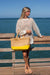 Person at beach holding the Lynwood natural raffia straw tote with linen lining and yellow color block base.