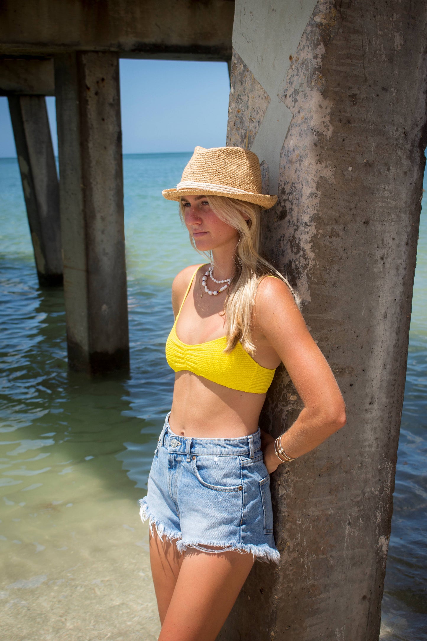 Person at the beach wearing the Anya & Niki Essential Hat - a natural colored crochet straw fedora hat with ivory rope band.