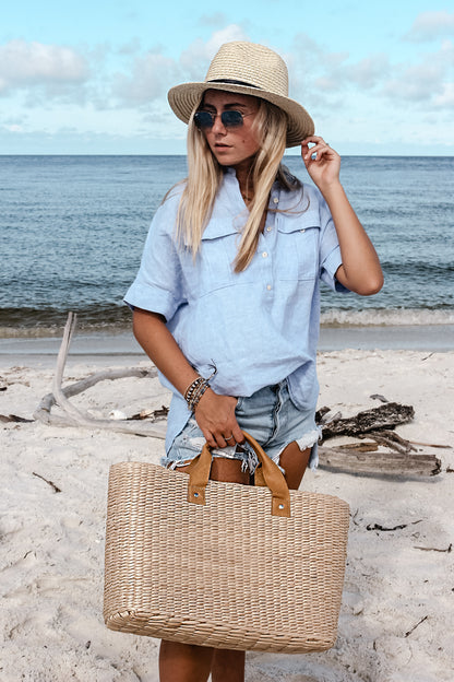 person at beach, holding large straw tote bag finished with natural leather handles and wearing straw fedora hat. 