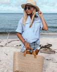 person at beach, holding large straw tote bag finished with natural leather handles and wearing straw fedora hat. 