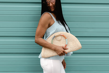 Person holding natural raffia straw half-moon clutch with rainbow colored wrapped circle handle and leather sides.