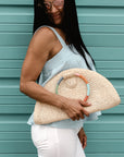 Person holding natural raffia straw half-moon clutch with rainbow colored wrapped circle handle and leather sides.