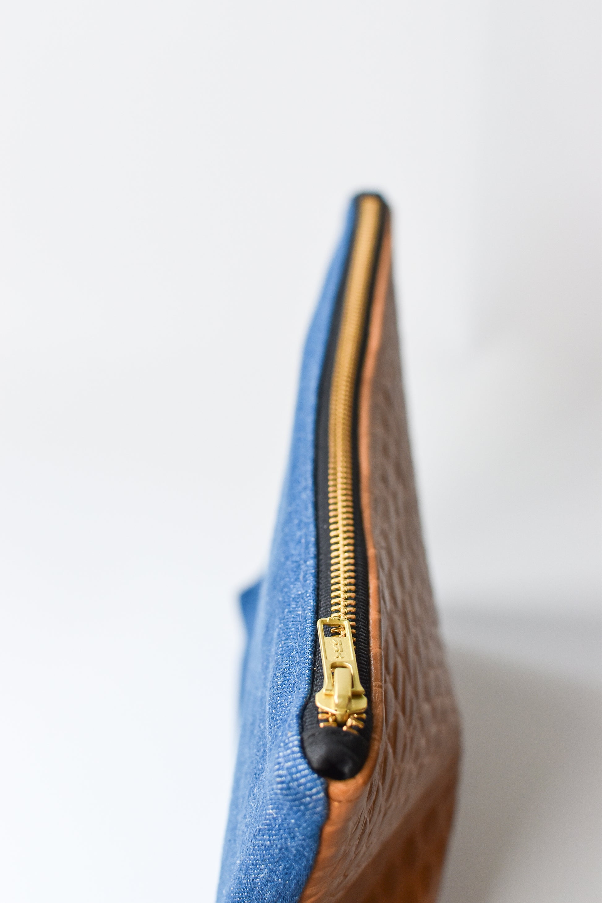 Medium wash denim and caramel colored embossed leather skin small pouch with brass zipper and leather logo label.