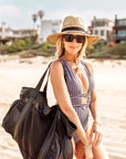 Person wearing straw hat with leather chin strap and holding XL black mesh beach bag